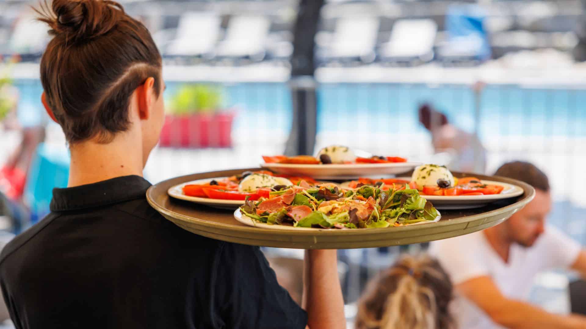 Présentation d'une Serveuse portant un plateau de salades fraîches dans un restaurant en plein air à Aluna Vacances