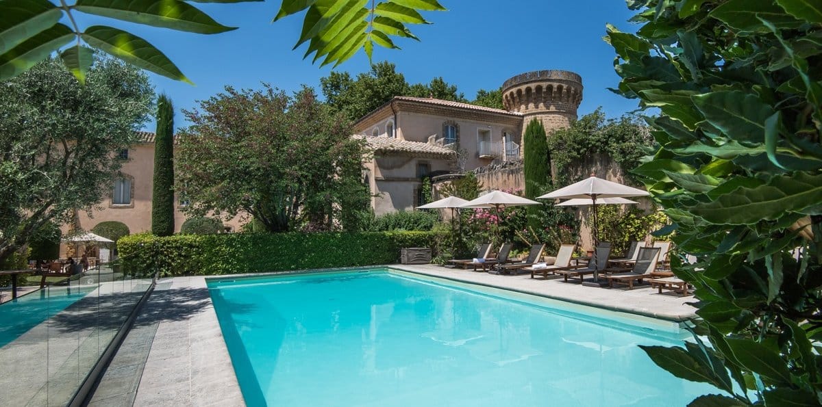 Piscine extérieure avec des chaises longues sous des parasols, entourée de jardins et une structure de château historique.