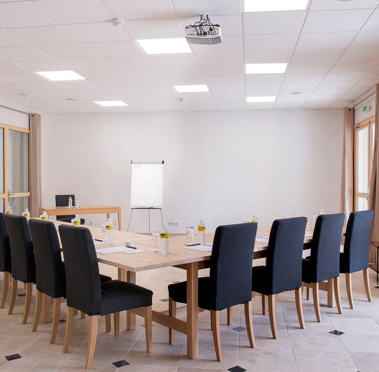 Salle de conférence moderne avec tables en bois, chaises noires, et un grand tableau blanc, sous un éclairage naturel.